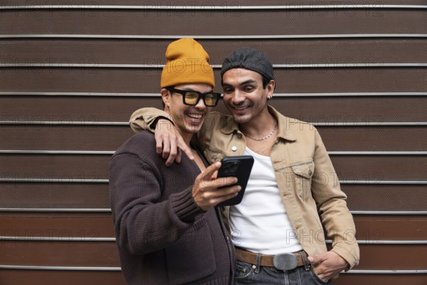 A Colombian gay couple smiles warmly as they look at a phone together. They are casually dressed, sharing a joyful moment outdoors in a relaxed setting