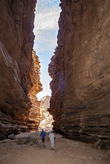 Two people walk through a narrow rocky gorge under a blue sky with clouds, rocky landscapes and gorges in the landscape of the Sahara in Algeria