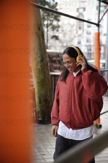 A man with long hair and glasses enjoys music through yellow headphones while walking outdoors in a red jacket, exuding happiness in an urban setting