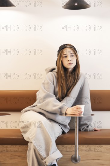 Teenage girl in casual attire with a cap, sitting thoughtfully on a bench. She holds a paper cup, relaxed and contemplative in a minimalist indoor setting