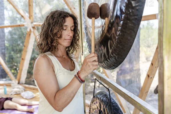 A female sound therapist plays a large gong during a gong bath session with eyes closed. The serene setting enhances relaxation and holistic wellness, offering a peaceful retreat experience