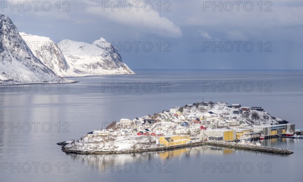 Scenic winter view of Husoy, Norway, with snow-covered houses nestled between serene waters and majestic mountains, creating a tranquil and picturesque coastal landscape