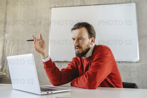 A office man in a red sweater gestures expressively during a virtual meeting. He is seated at a desk with a laptop and whiteboard, embodying remote work dynamics