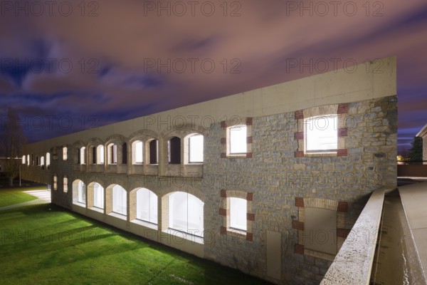 A beautifully illuminated stone hospital with arches under a twilight sky, showcasing architectural elegance. Green grass contrasts with the vivid sky, capturing serenity