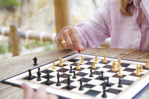 A close-up of hands moving chess pieces during a game played outside on a wooden table The board displays a strategic mid-game position on a sunny day