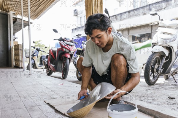 A Balinese artist skillfully paints a sculpture outdoors, blending traditional techniques with natural surroundings. Vibrant motorcycles are seen in the background, adding a modern touch