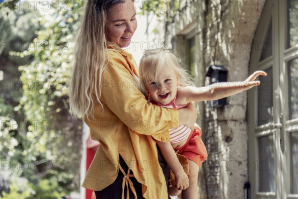 A joyful mother lovingly holds her daughter in an outdoor setting, surrounded by nature. The scene captures a tender moment, showcasing the bond between parent and child