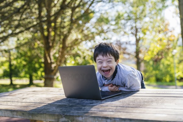 A cheerful child with Down syndrome is smiling joyfully at a laptop while sitting at a wooden table in a sunlit park. The image conveys happiness and technology