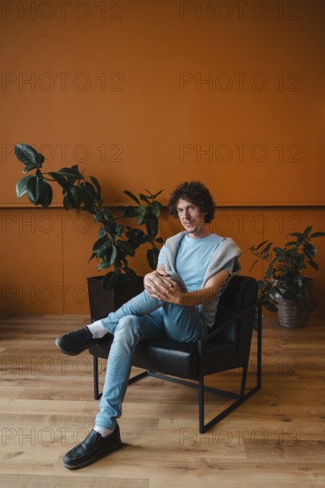 A young man with curly hair sits in a stylish chair in a contemporary room. The space features a warm, orange wall and lush green plants, creating a relaxing atmosphere