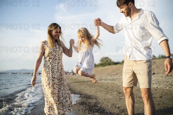 A cheerful family enjoys their time by the sea in Italy, as the parents swing their daughter into the air while walking along the shoreline. The child laughs with excitement and joy