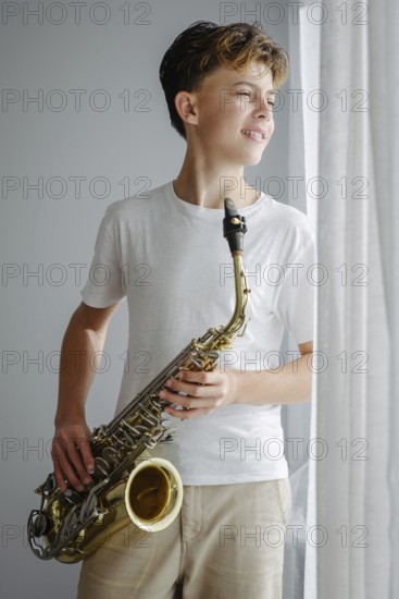 A young saxophonist in a white shirt looks out the window, standing in soft sunlight. The relaxed pose captures a moment of inspiration and musical passion