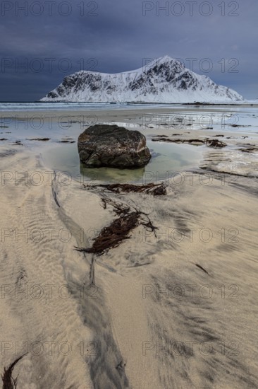 Rocks lying on the beach in front of snowy mountains, dark clouds, winter, Flakstadoya, Lofoten, Norway