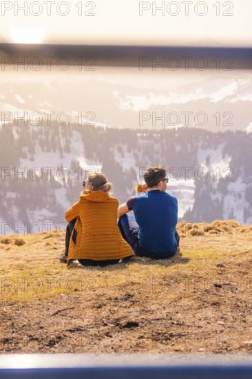 Couple sitting on a mountain peak with a view of the sunny winter valley, Hochgrat Mountains, Allgäu Alps, Oberreute, Germany