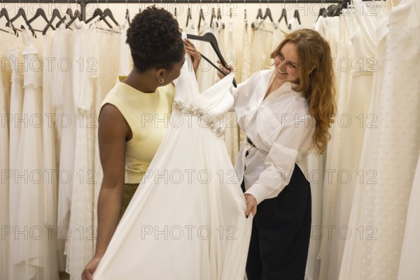 Two women joyfully examining a wedding dress in a bridal shop, surrounded by elegant gowns. The scene captures the excitement and anticipation of wedding preparations