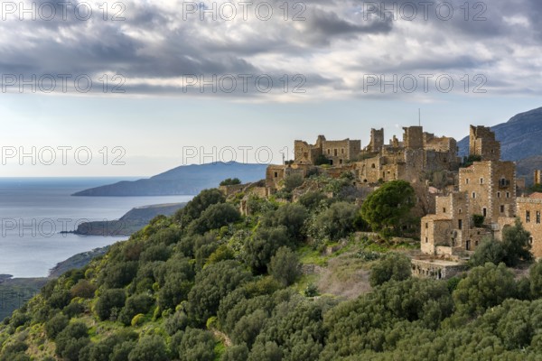 A view of Vathia, a historic abandoned village in Mani Peninsula, Greece, with traditional stone towers perched on a hillside overlooking the sea and surrounded by lush greenery