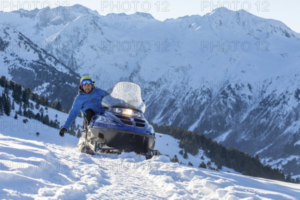 A rider on a snowmobile speeds through a snow-covered mountainous landscape, capturing the thrill of winter sports against a backdrop of majestic peaks and clear blue skies