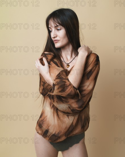 Studio portrait of a young woman posing with a thoughtful expression, wearing a patterned blouse and fishnet stockings against a beige background