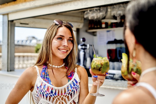 Two friends are at a vibrant beach bar during a sunny summer day, holding colorful cocktails and sharing smiles. The atmosphere is relaxed and filled with joy