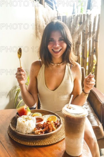 Young woman holding cutlery and smiling, ready to enjoy traditional filipino breakfast with iced coffee in a restaurant in siargao island, philippines