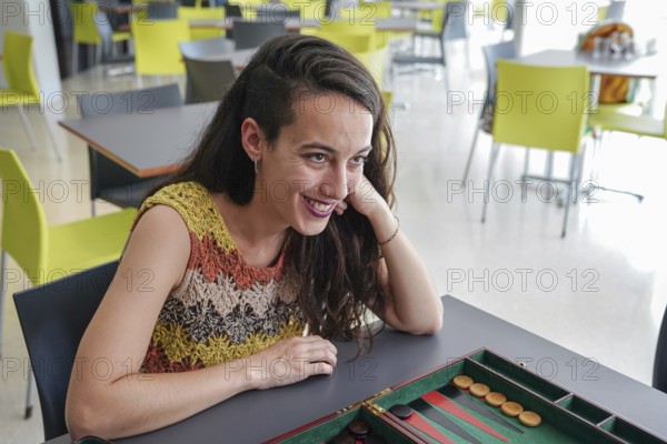 A woman sits at a table in a modern cafe, smiling and engaged, while playing a game of backgammon Bright chairs and natural light create a lively atmosphere