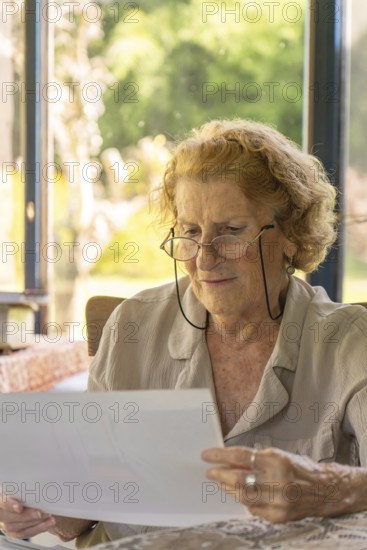 Elderly woman with glasses reading documents at home, sitting at a table with natural light coming through a window, enjoying a peaceful moment