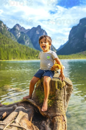 Carefree child sticking out tongue, sitting on tree stump by lake dobbiaco with plush toy, enjoying summer vacation in the italian dolomites