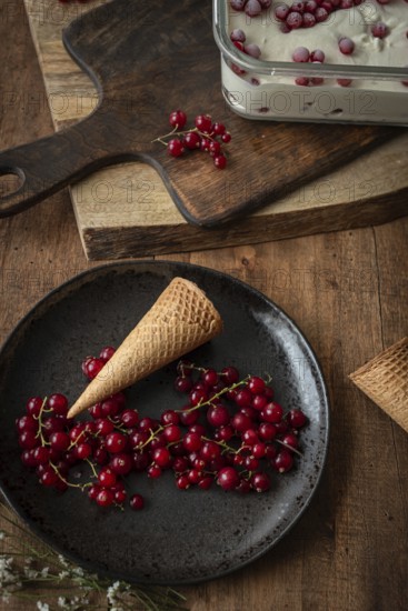 Top view of a rustic food scene featuring red currants on a dark plate beside a waffle cone, set against a backdrop of a wooden board and a glass dish filled with ice cream sprinkled with berries