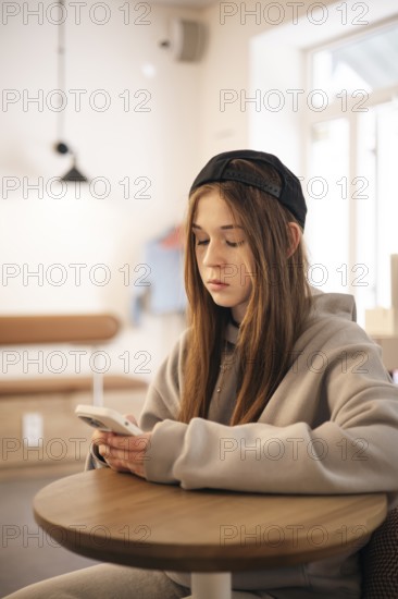 A teenage girl wearing a hoodie and cap sits at a small table, engrossed in her smartphone. The warm, softly-lit environment suggests a cozy, relaxed atmosphere