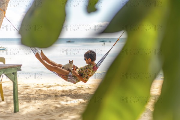 A woman and a cat enjoy a serene moment in a hammock on a sandy beach, surrounded by lush green foliage under bright sunlight, exemplifying a tranquil vacation scene in the Philippines