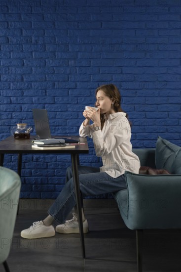 A woman in casual attire sips tea, seated at a table with a laptop, against a vibrant blue brick wall. The modern setting exudes a relaxed yet productive atmosphere