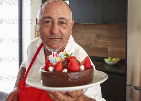 A cheerful senior man in a red apron holds a chocolate cake adorned with strawberries and a Happy Birthday decoration, celebrating his 50th birthday with a warm smile