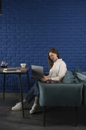 A woman in casual attire works on a laptop, seated on a teal sofa against a blue brick wall. A table with tea and notebooks complements the cozy atmosphere