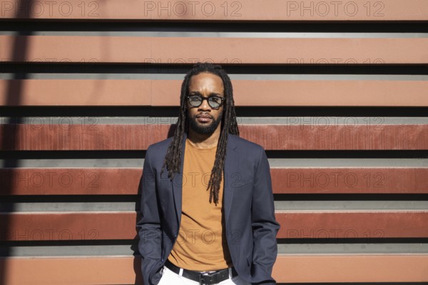 Stylish cuban business man with dreadlocks in sunglasses and casual blazer stands against modern striped wall under sunlight, exuding confidence and urban elegance