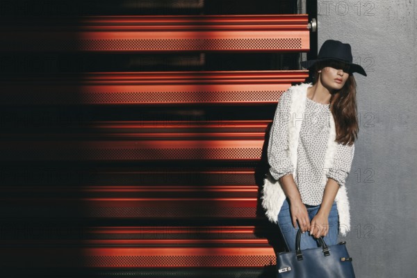 A fashionable woman in a wide-brimmed hat and stylish clothing poses with a handbag against a red background, casting dramatic shadows and exuding urban chic