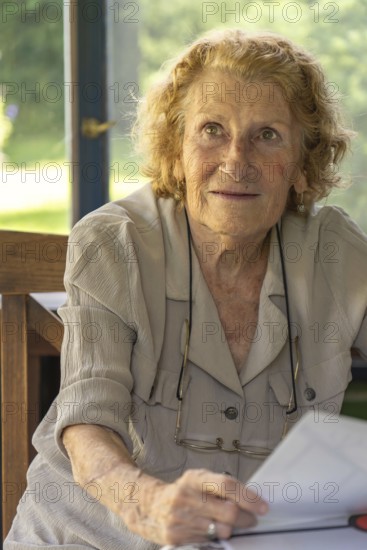Portrait of a senior woman sitting at a table on a sunny veranda, holding and reading a book, embracing the joys of retirement with a peaceful and content smile