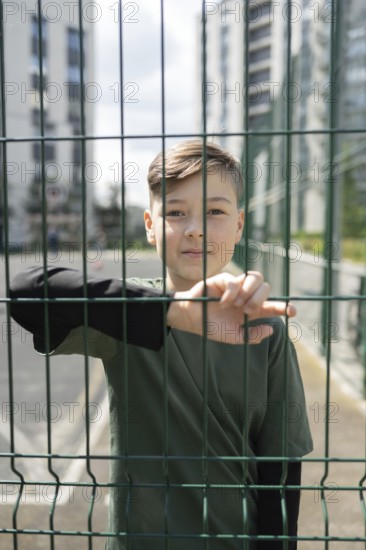 A young boy in a dark shirt stands pensively behind a green wire fence, with urban high-rise buildings blurred in the background, conveying a sense of reflection and focus