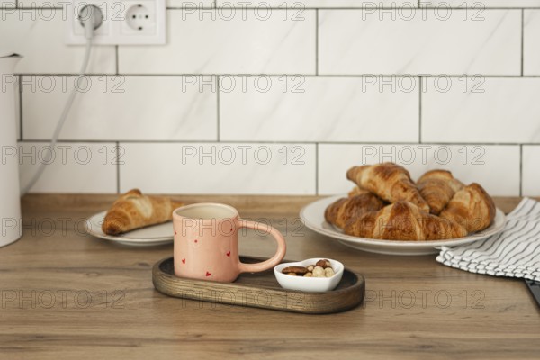 Freshly baked homemade croissants served on a wooden table with a cozy pink mug and a small bowl of nuts. A perfect start to a relaxing morning at home