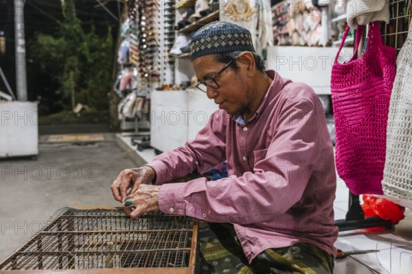 A balinese artisan focuses intently while crafting at a market stall. Surrounded by handmade merchandise, he sits engrossed in his work, showcasing traditional craftsmanship