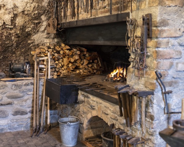 A rustic blacksmith workshop featuring a burning forge, neatly stacked firewood, and an array of traditional tools. The stone and brick setting highlights old-world craftsmanship