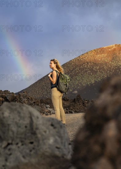 A woman with a backpack explores the volcanic landscape of Timanfaya National Park in Lanzarote. A vivid rainbow arches across the dramatic sky, highlighting the natural beauty