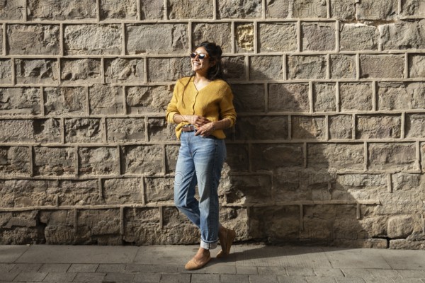 A Latin woman in casual attire, wearing sunglasses, smiles while leaning against a sunlit brick wall. Her relaxed pose and cheerful expression convey a sense of joy