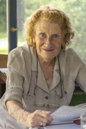 Happy retired woman with curly hair holding a book and smiling, enjoying her leisure time at home, sitting at a table by the window with eyeglasses hanging from her shirt