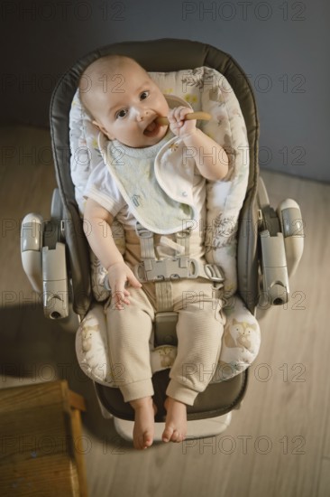 A baby is sitting in a high chair, holding a spoon and looking at something. The room has soft lighting. The baby wears a bib and light pants. A wooden table is nearby