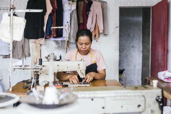 A asian seamstress works on a sewing machine in a cozy workshop. Surrounded by hanging clothes and sewing tools, she displays craftsmanship and concentration