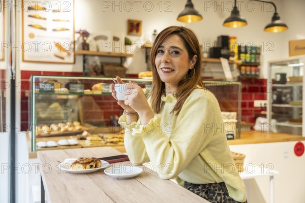 Young woman in a light yellow sweater smiles at a sunny cafe counter, holding espresso and enjoying a fresh pastry amid warm, modern bakery decor and natural window light
