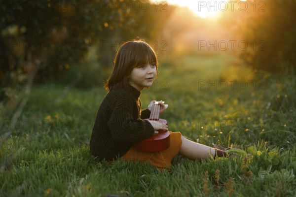 A young child sits on lush grass with orange trees, playing a red ukulele in a sunlit meadow. The warm glow of sunset surrounds the peaceful scene, evoking joy and tranquility
