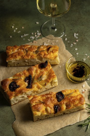 Freshly baked homemade focaccia topped with garlic and olives served on parchment paper. Accompanied by olive oil dip and a glass of white wine on a textured green surface