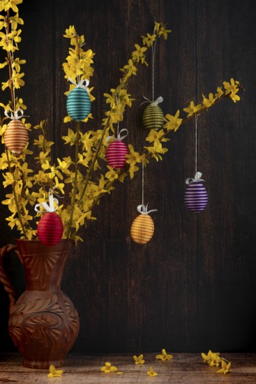 Branches of forsythia (Forsythia) in a rustic jug hung with Easter eggs against a dark background