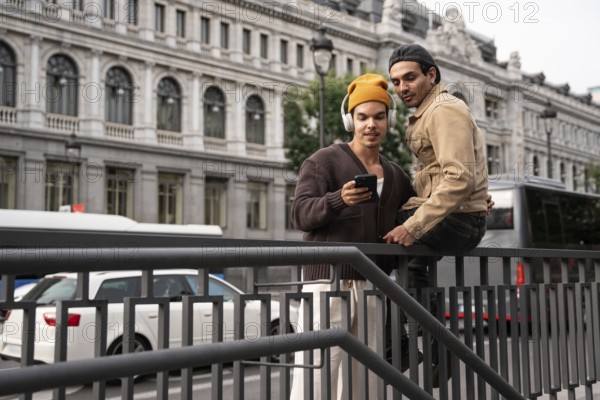 A gay couple in casual attire shares a tender moment on a Madrid street, leaning on a railing and looking at a smartphone. They convey a sense of connection and modern romance