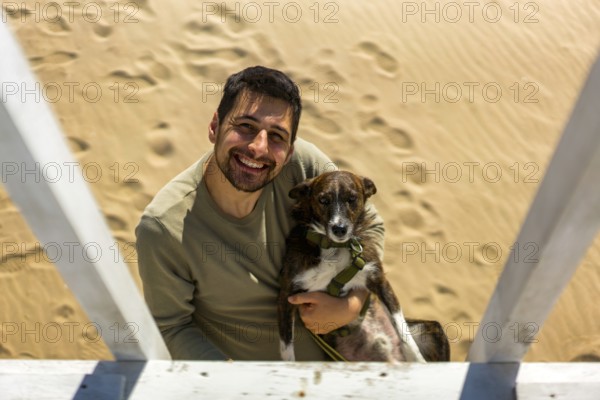A cheerful man holding his dog on a sunny beach in Greece. The sand is marked with footprints. Captures joy and companionship in a serene coastal setting